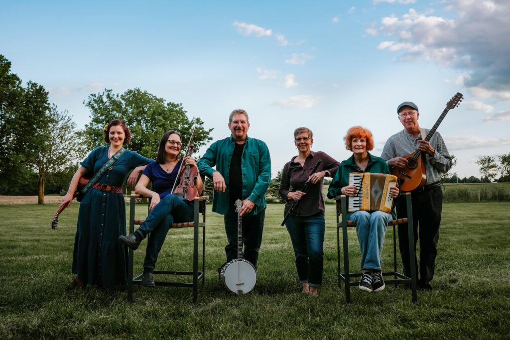 six musicians standing and sitting in a row in a field, posing, holding guitar, fiddle, banjo, flute, accordion, and bouzouki