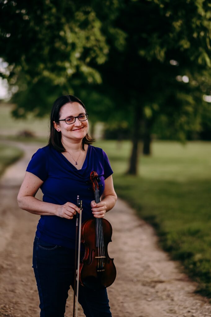 Sarah Riskind standing and smiling on a dirt road, holding violin and bow at her side