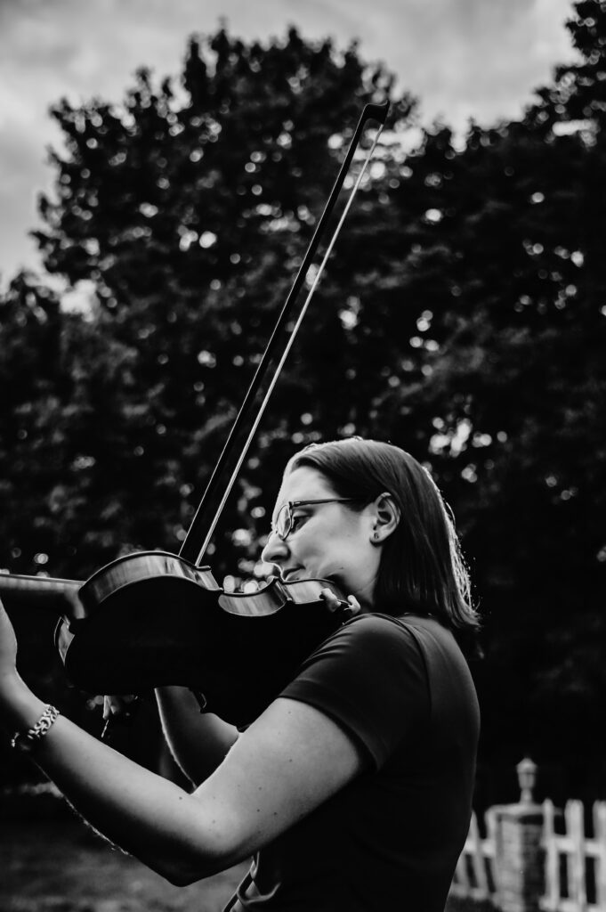 Black and white photo Sarah Riskind playing the violin in front of trees and a fence